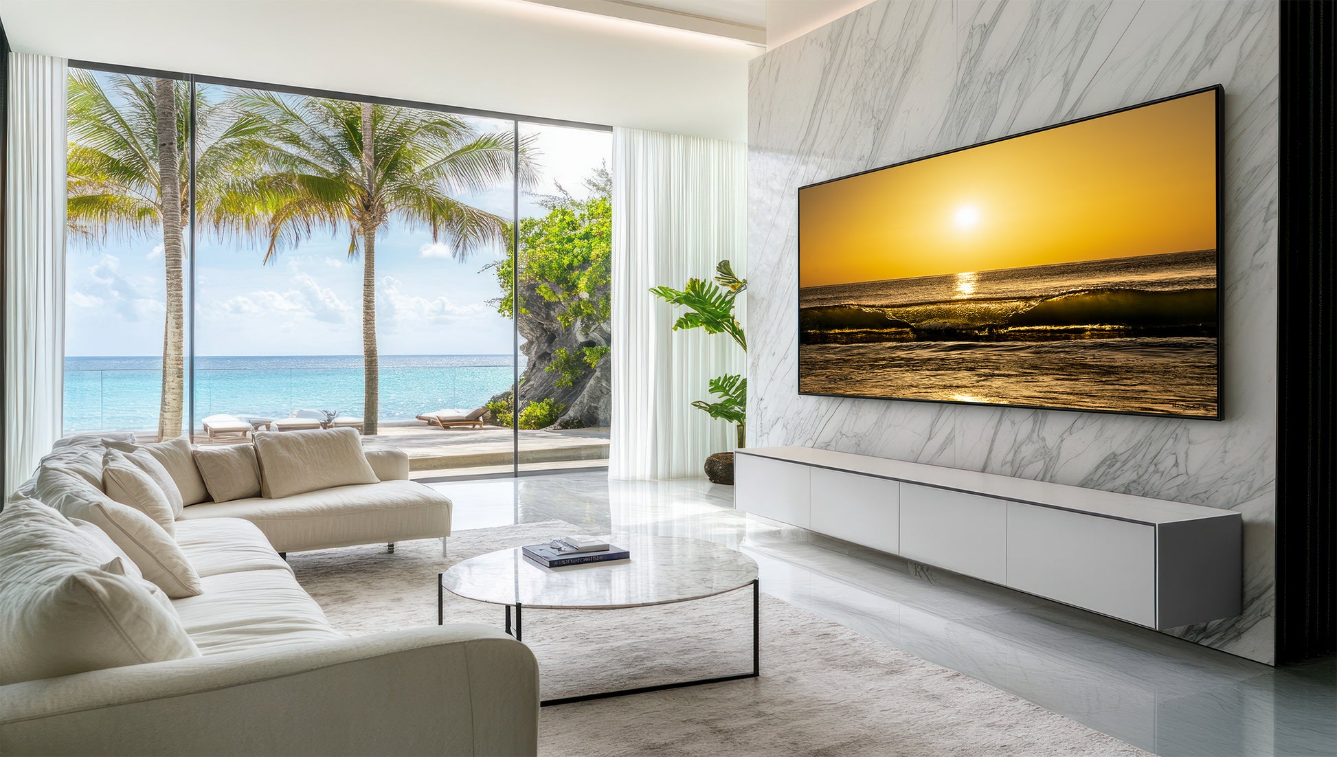 A large framed wall art print depicting a golden sunrise over ocean waves, displayed in a modern living room with a white sectional sofa, glass coffee table, and a large window overlooking a tropical beach with palm trees.