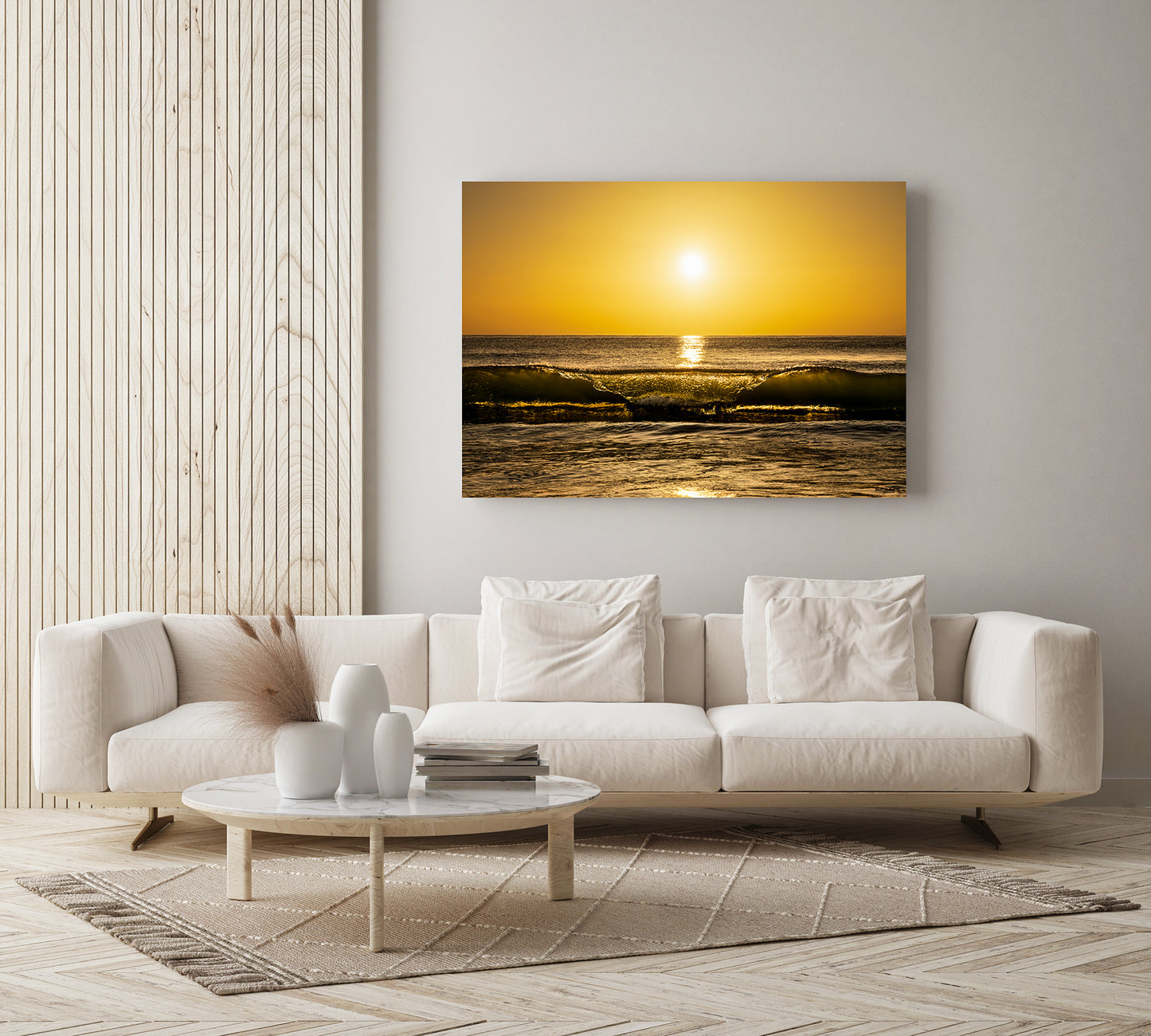 A rectangular wall art print showing a golden sunrise over ocean waves, displayed above a white sofa in a modern living room with a round coffee table and light wood flooring.